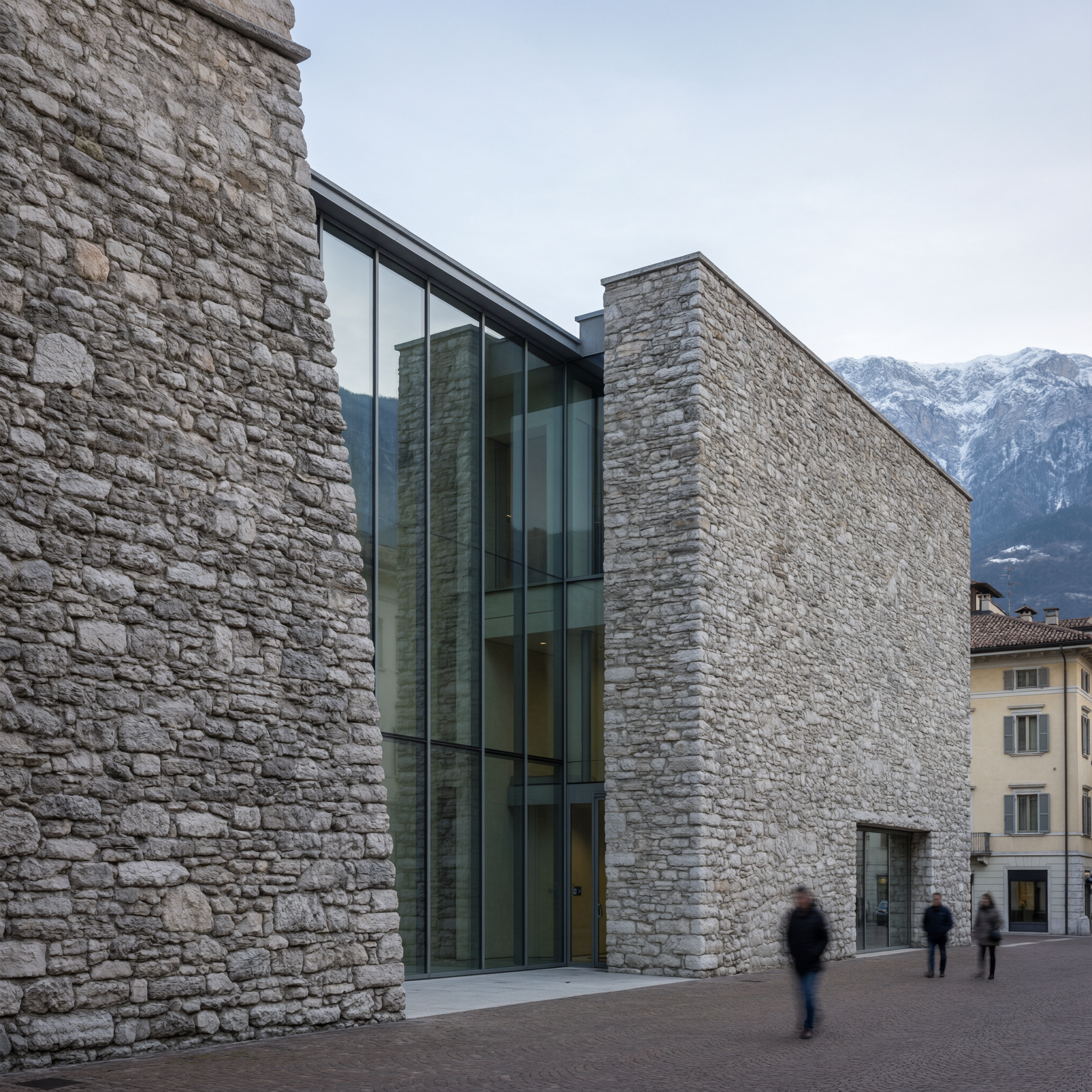 Modern stone building with tall glass entrance doors in Trento, with snow-capped mountains visible in background